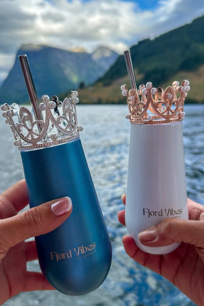 bilde av to hender som holder hver sin prosecco-kopp i hhv hvit og blå med her sin pyntekrone og sugerør. De skåler foran fjord og fjell