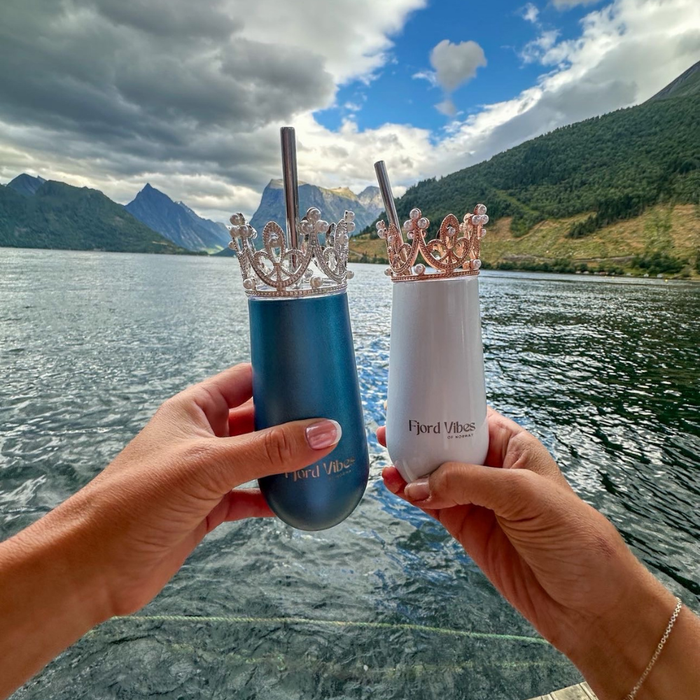 bilde av to hender som holder hver sin prosecco-kopp i hhv hvit og blå med her sin pyntekrone og sugerør. De skåler foran fjord og fjell