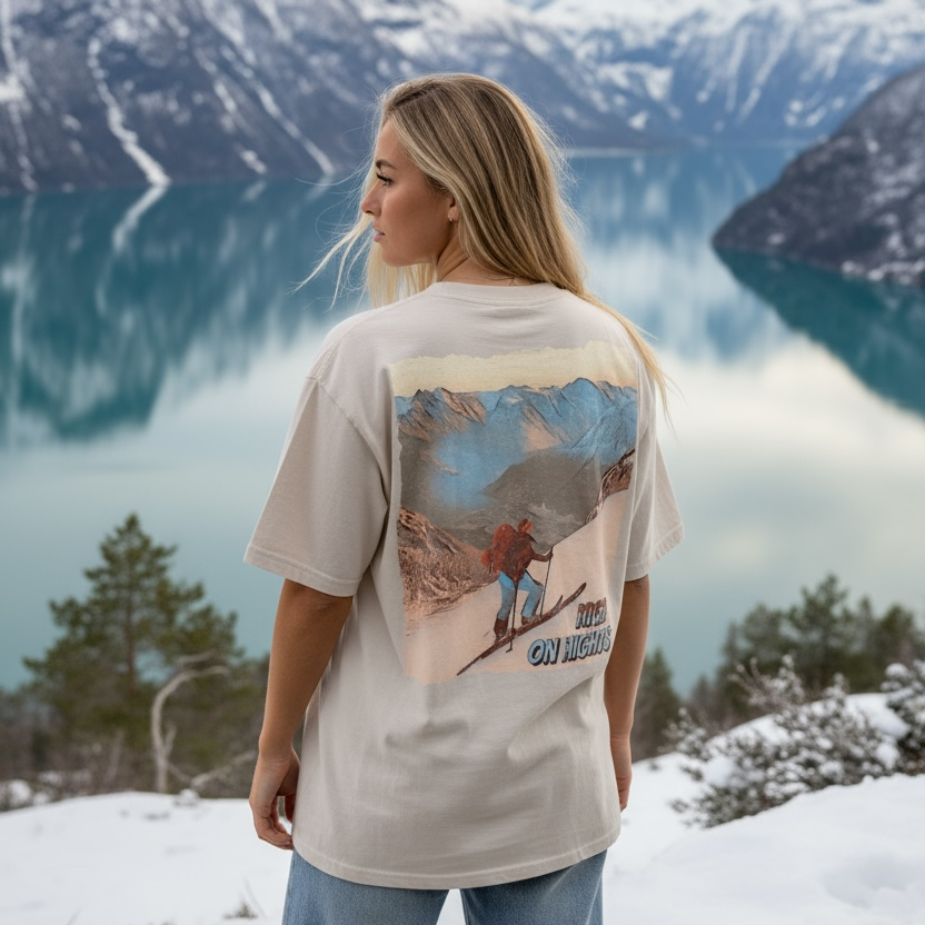 Dame som har på en oversized beige washed tee med ski-motiv trykka på hele ryggen. Hun står ved en fjord med snø på fjelltoppene