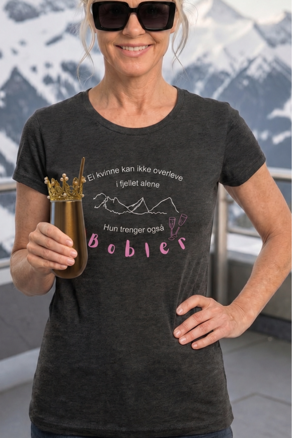 Prosecco t-skjorte i melert mørk grå farge med hvit og rosa skrift, dame t-skjorte modell med påskrift ei dame kan ikkje overleva i fjellet åleine, ho treng også bobler. En dam e som står på en terrasse i fjellheimen og holder en termokopp med bobler i hendene