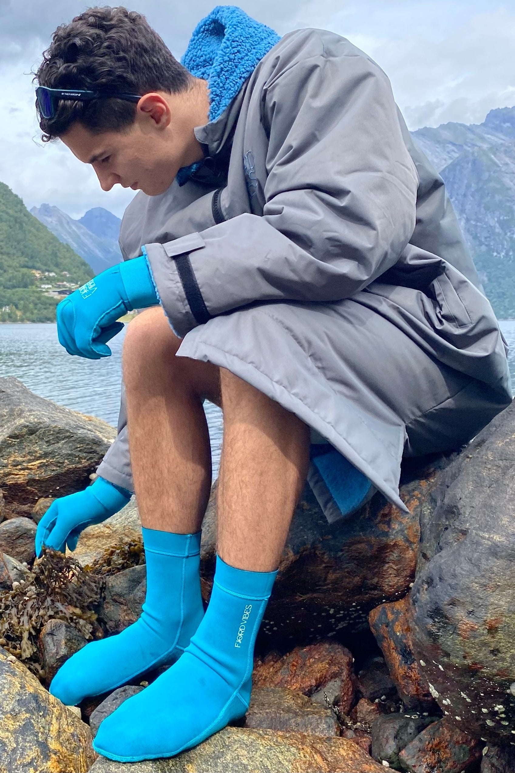 Salty but Sweet™ Polar Fjord Robe ECO, Glacier-blue surf-robe - DoUdare - badeponcho - blå - unisex En mann som sitter på en strand og har på en grå tykk poncho laget for isbading