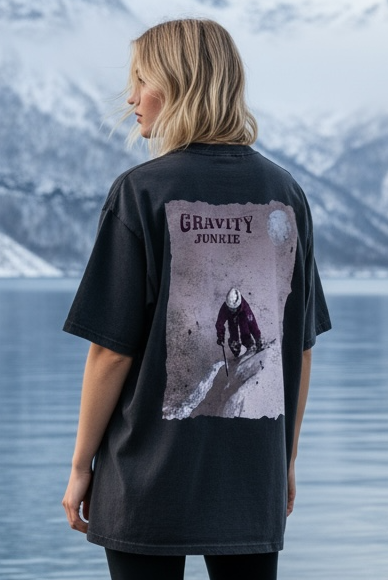 Dame som har på en oversized grå washed tee med ski-motiv trykka på hele ryggen. Hun står ved en fjord med snø på fjelltoppene