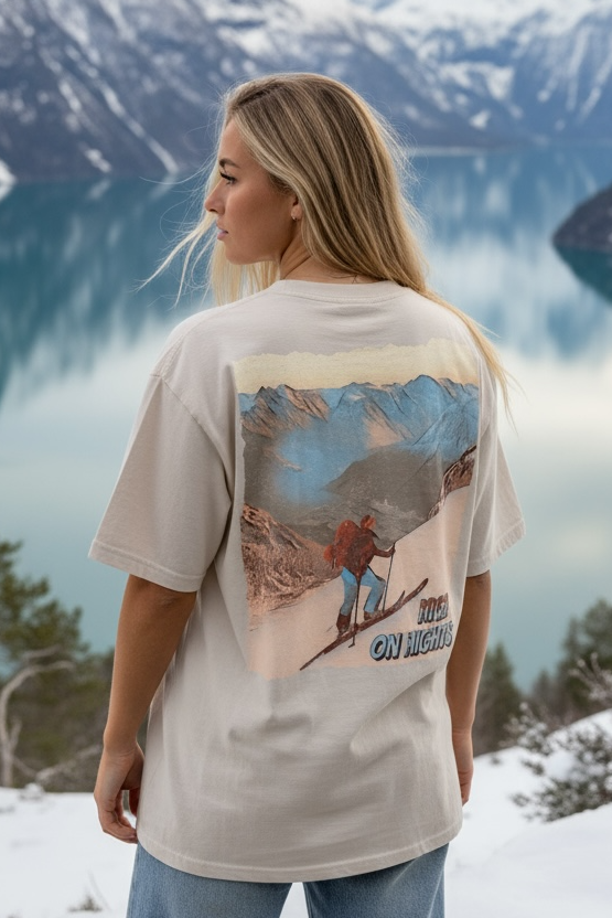 Dame som har på en oversized beige washed tee med ski-motiv trykka på hele ryggen. Hun står ved en fjord med snø på fjelltoppene